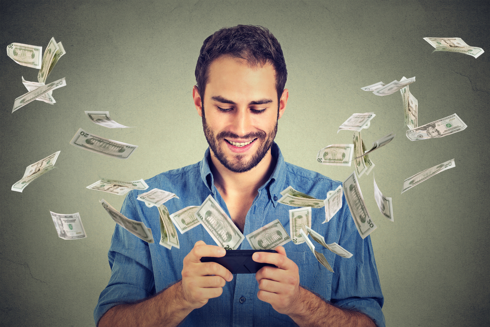 Technology online banking money transfer, e-commerce concept. Happy young man using smartphone with dollar bills flying away from screen isolated on gray wall office background.